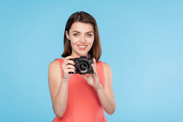 Happy young woman with toothy smile holding camera in front of her while going to take photo of you