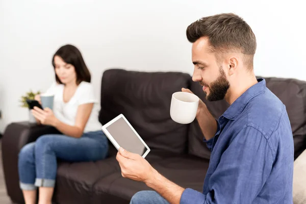 カジュアルなシャツを着た好奇心旺盛な若者がソファに座り ガールフレンドと一緒にスマートフォンに夢中になり コーヒーを飲みながらタブレットでオンラインニュースを読む — ストック写真