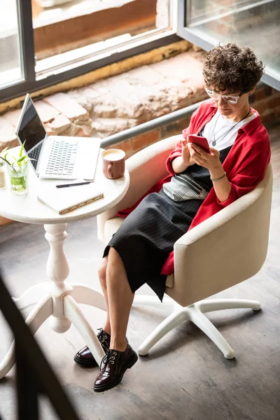 Young mobile woman with smartphone messaging while sitting in armchair by table in office or cafe
