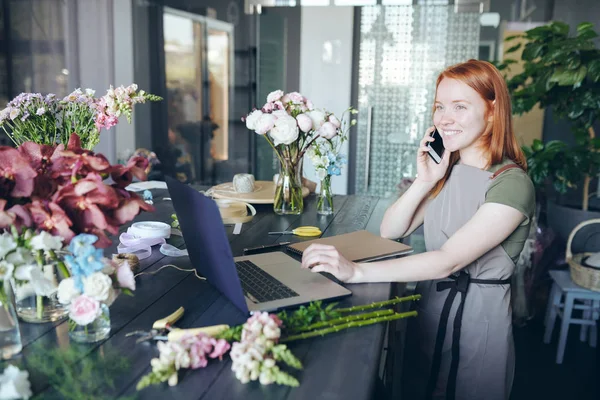 様々な花でいっぱいのカウンターに立ち 電話で注文を話しながらノートパソコンを使用して 花屋のポジティブな若いマネージャー — ストック写真