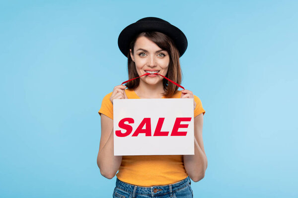 Pretty young woman in hat announcing seasonal sale while holding paper with red word and looking at you