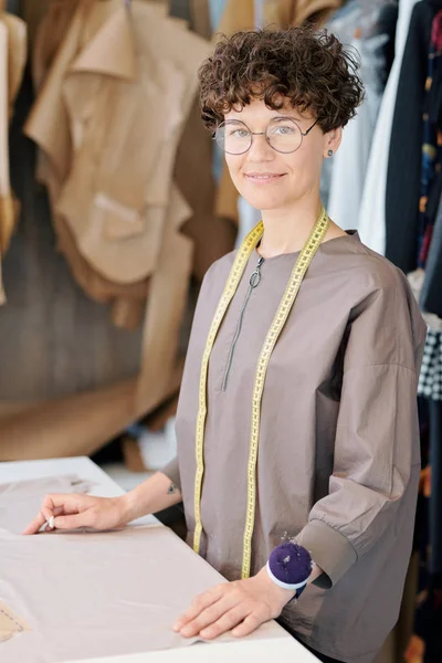 年轻的女性裁缝与测量磁带看着你 而站在办公桌与一块织物 — 图库照片