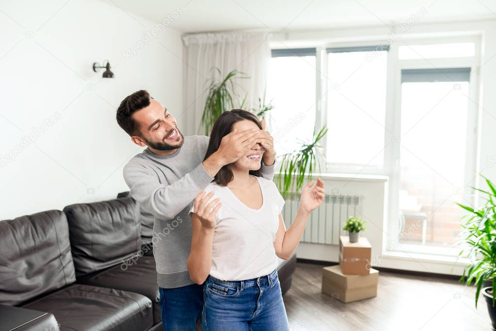 Happy young couple in casual clothing standing in living room with moving boxes, bearded man covering eyes to girlfriend
