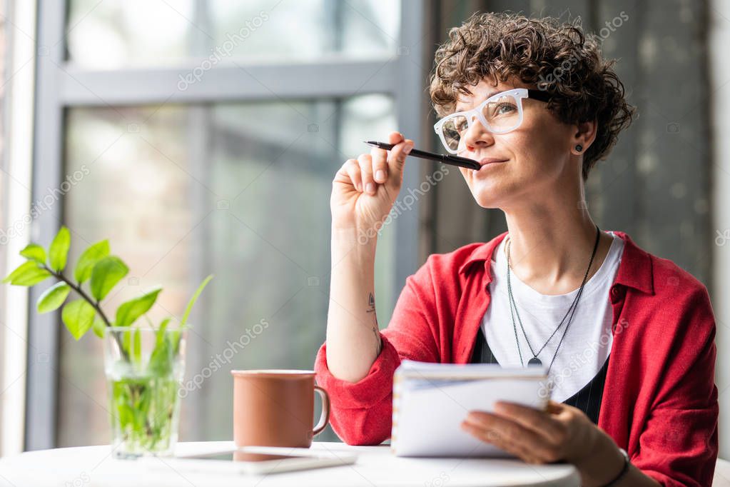 Young pensive female in eyeglasses thinking of new creative ideas while sitting by table in cafe or office