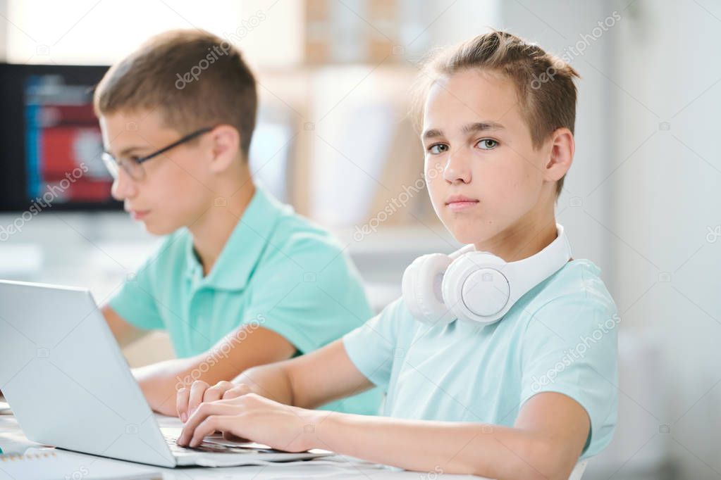 Cute and clever schoolboy with headpones on neck working over homework or presentation in front of laptop