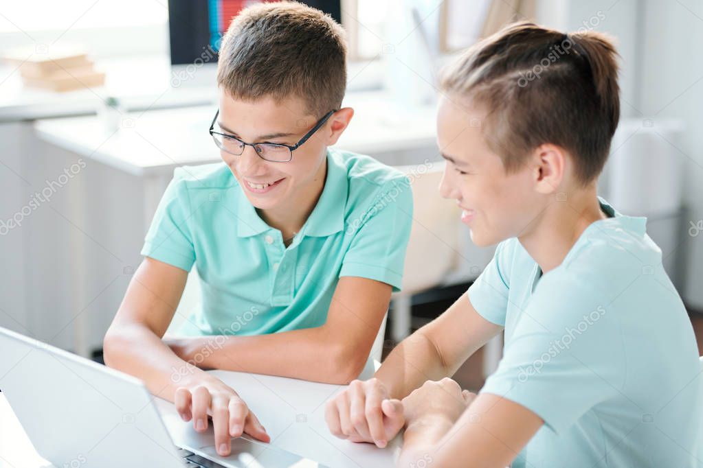Two cute middle school boys sitting by desk in front of laptop display and preparing home assignment
