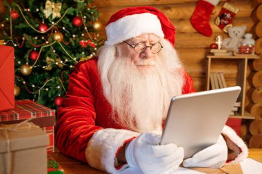 Ciddi Noel Baba evde, tahta masada oturuyor. Önünde tablet var ve internetten Noel dilekleri süzülüyor.