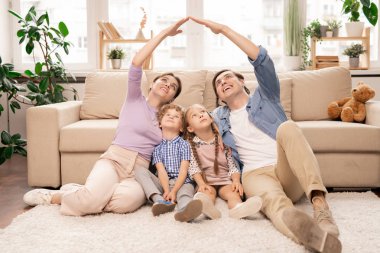 Genç ebeveynler ellerini birbirine yakın tutarak oturma odasının zemininde otururken iki küçük çocuklarının başını sokacakları bir çatı oluşturuyorlar.