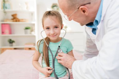 Steteskopla gülümseyen küçük bir kız kalp atışlarını dinlerken doktor göğsünde tıbbi aleti tutuyor.