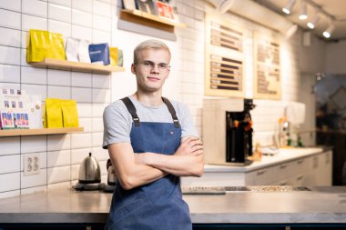 Genç çapraz kollu barista ya da kafeterya sahibi çalışma ekipmanları ve kahve paketlerinin arka planında duruyor.