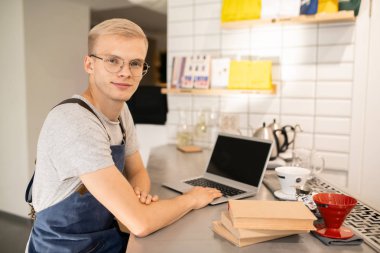 Üniformalı ve gözlüklü genç adam iş yerinde duruyor ve dizüstü bilgisayarın önünde öğrenirken sana bakıyor.