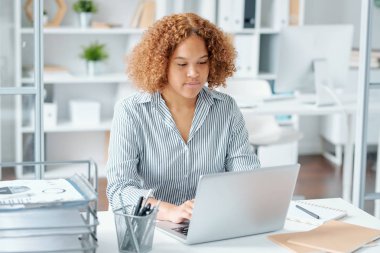 Genç melez iş kadını dizüstü bilgisayarın önünde oturmuş ofisteki çevrimiçi verileri yazıyor ve inceliyor.
