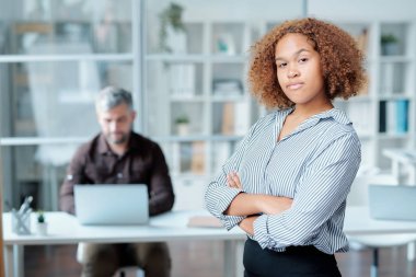 Genç kendine güvenen, kıvırcık saçlı, kollarını göğsüne dolayıp kameranın önünde duran ve sana bakan ofis müdürü.