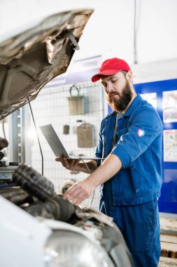 Genç sakallı tamirci bozuk kamyon motorunu inceliyor ve internette bilgi bulmak için dizüstü bilgisayar kullanıyor.