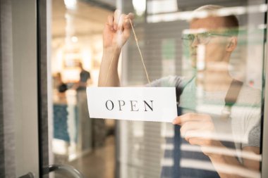 Kafeteryanın şeffaf kapısında duran ve kafenin açık olduğunu söyleyen kağıt uyarı asan genç barista.