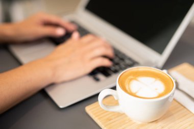 Kafedeki şebekede bilgisayar tuşlarına dokunan kız öğrencinin elindeki ahşap tepside bir fincan cappuccino.