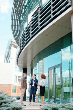 Üç genç zarif iş ortağı çalışma saatlerini tartışırken modern ofis binasının yanında duruyorlar.