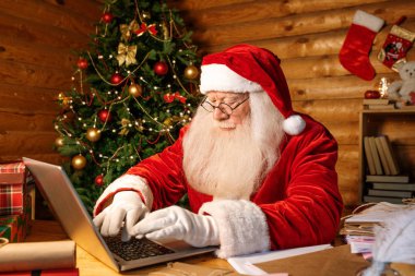 Çağdaş Noel Baba, masa başında oturup Noel hediyesi almak için çevrimiçi dükkanlar ararken dizüstü bilgisayarında anahtarlar yazıyor.