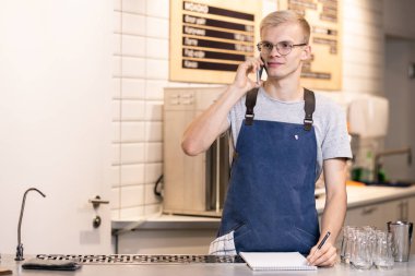 Önlüklü ve tişörtlü genç barista iş yerinde durup not defterine not alarak müşterilerinin siparişlerini alıyor.