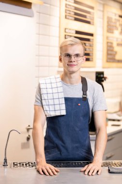 Dost canlısı genç sarışın erkek barista, omuzlarında havluyla iş yerinde kameranın önünde duruyor ve sana bakıyor.