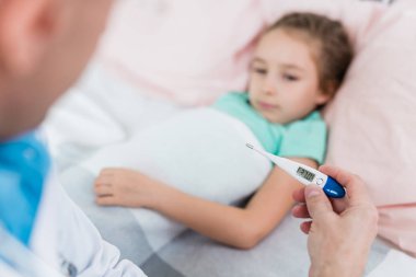 Hasta küçük bir kızın evdeki yatağında yatan çocuk doktorunun elinde tıbbi termometre.