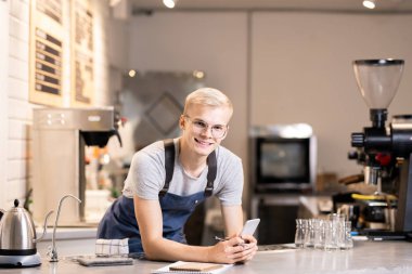 İş kıyafetleri içinde genç neşeli barista masanın üzerine eğilip akıllı telefondan müşterilerin siparişlerini karıştırırken sana bakıyor.