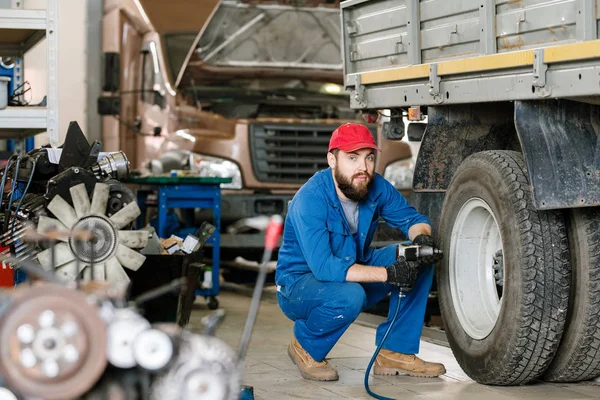 穿着工作服的有自信的技师坐在卡车轮旁修理时看着你 — 图库照片