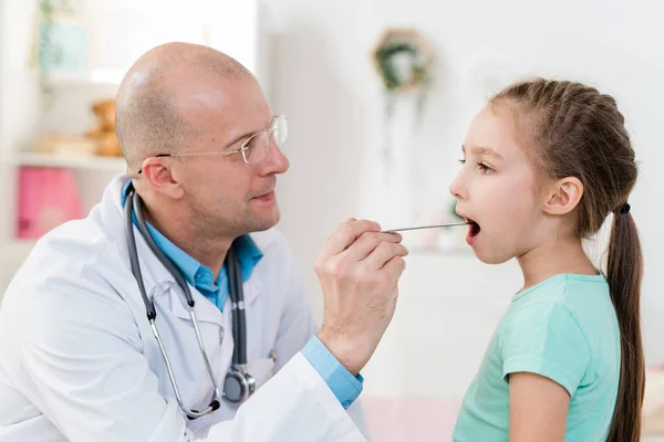 生病的小女孩张大嘴 而医生则用医疗仪器检查她的喉头痛 — 图库照片