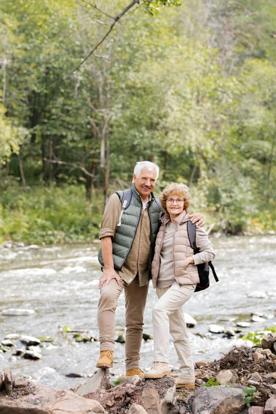 周末旅行时 两个快乐的老徒步旅行者背着背包站在河边看着你 — 图库照片
