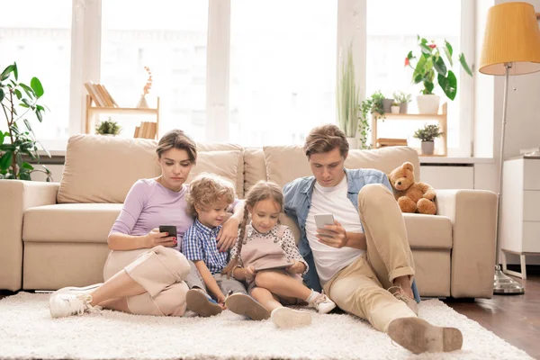 由一对年轻夫妇和两个孩子组成的现代家庭 他们坐在沙发旁边的地板上 在客厅里用他们的移动小玩意 — 图库照片