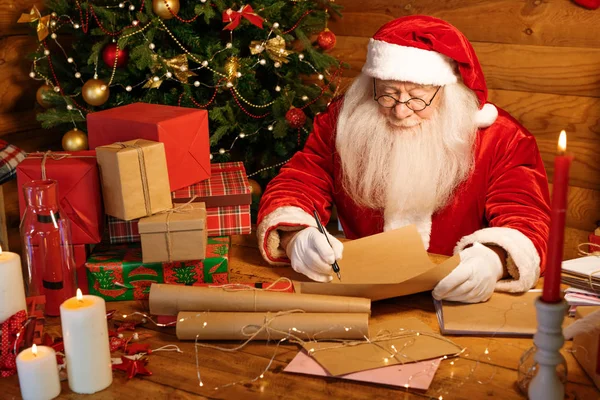 白い長い髭を生やしたサンタは 木製のテーブルに座って装飾された木で座り クリスマスの手紙のために答えを書きます — ストック写真