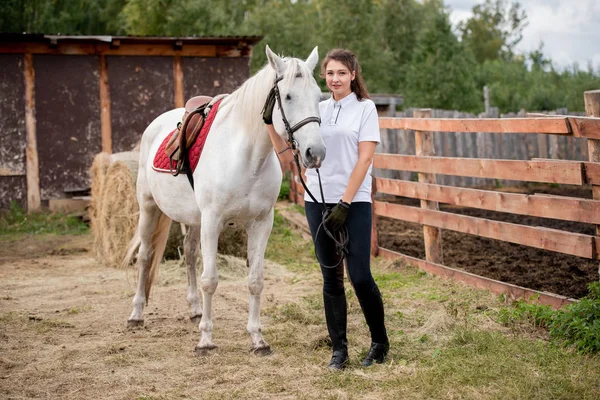 若いですアクティブ女性でジーンズ ポロシャツとブーツ見ている間にあなたを冷却しますとともに白い純血種競走馬で農村環境 — ストック写真