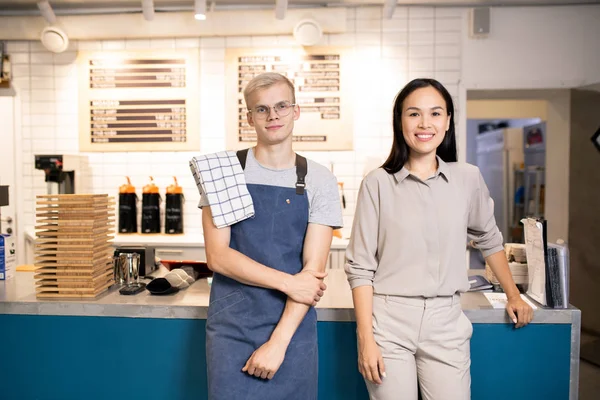 Two young best waiters of luxurious restaurant or cafe standing by counter while meeting new guests