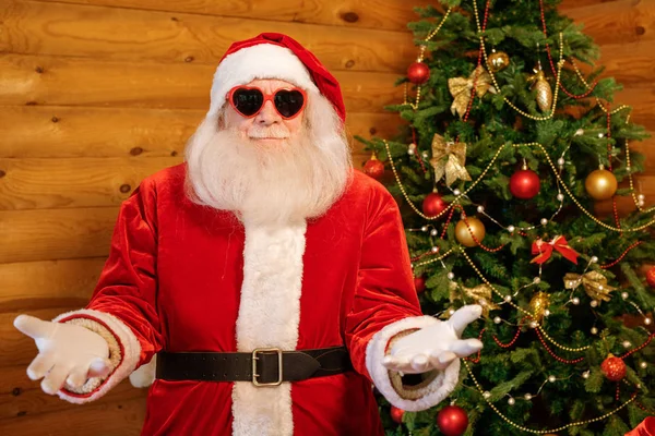 快乐迷人的圣诞老人 身穿服装 戴着心形太阳镜 站在镜头前 背景装饰着圣诞树 — 图库照片