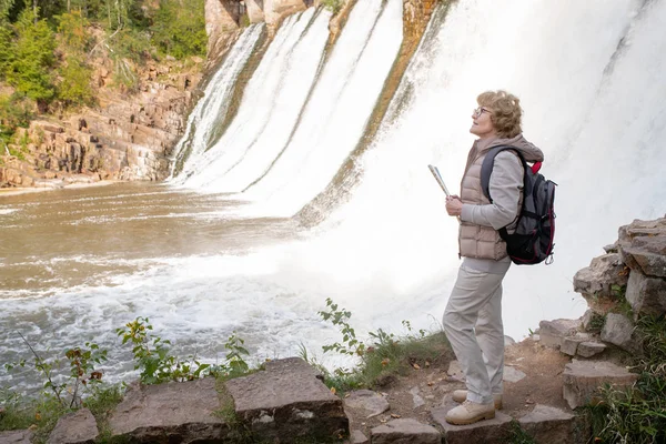 带背包和地图指南的成熟女性徒步旅行者站在自然环境中的瀑布边 寻找正确的道路 — 图库照片