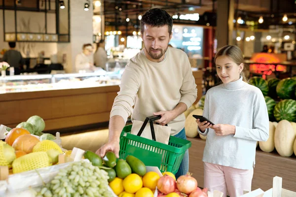 在参观大型现代超市时 提篮子的年轻人和他的女儿选择鳄梨和其他水果 — 图库照片
