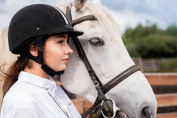 田舎の環境で競走馬のそばに立つ白いシャツと乗馬ヘルメットの若い穏やかな女性 — ストック写真