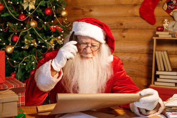 圣诞前带着手机给父母打电话的流动圣诞老人 一边翻阅着他们孩子的名字 一边在纸上写着 — 图库照片