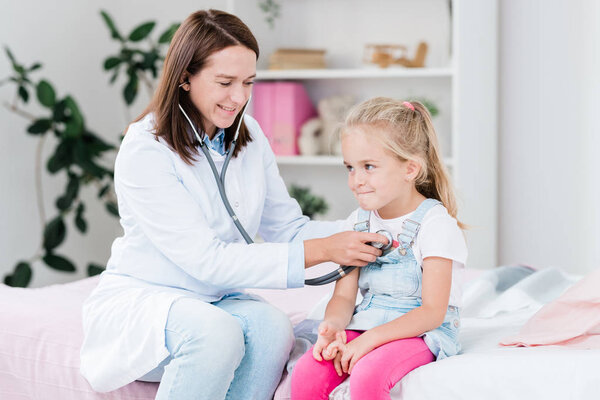 Sick little girl sitting on bed in medical office while pretty doctor examining her chest with stethoscope