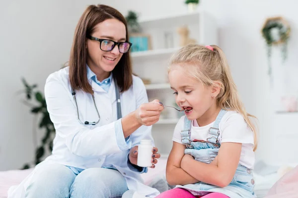 Sick little girl taking medicine from spoon held by young female doctor in whitecoat while visiting hospital