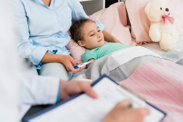 Niña Enferma Acostada Cama Debajo Manta Mientras Madre Sostiene Termómetro — Foto de Stock