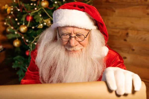 当代的圣诞老人穿着服装 戴着眼镜 手里拿着写着圣诞祝福的纸看书 — 图库照片
