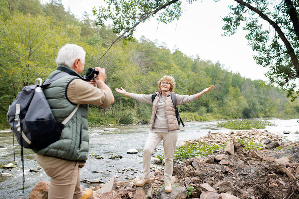 Cheerful aged active woman with outstretched arms looking at her husband with camera while both standing by river