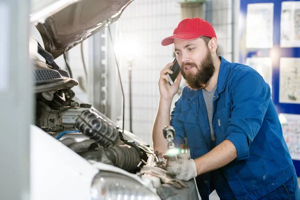 带胡子的机器维修工站在坏汽车的发动机旁 一边用手机与客户交谈 — 图库照片