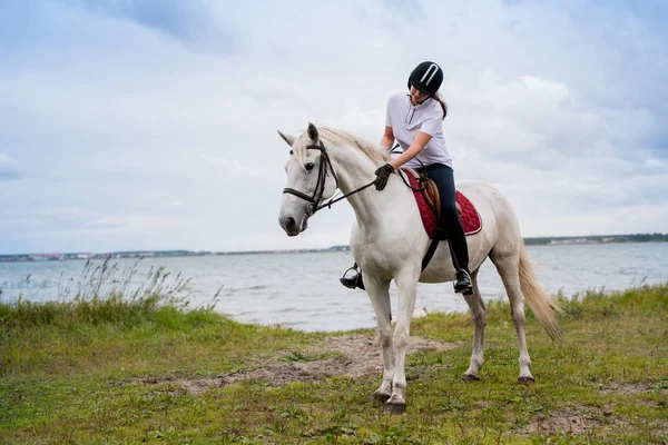 白い競走馬の後ろに座りながら川沿いに動く乗馬姿の若い現役女性 — ストック写真