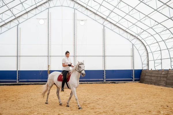 乗馬センター内の砂のアリーナを下に移動しながら馬の後ろに座って若いアクティブな女性 — ストック写真