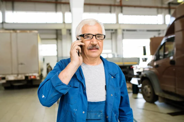 Senior professional technician in workwear and eyeglasses standing in workshop and consulting client on mobile phone