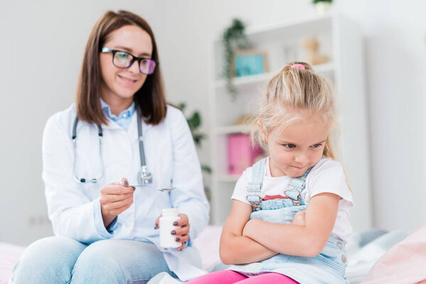 Naughty sick little girl turning away from doctor giving her medicine while both sitting in medical office