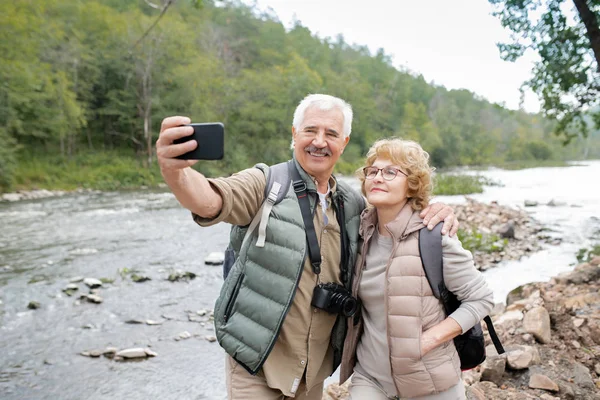 旅途中 在森林河边自拍时 快乐而成熟的活跃配偶看着智能手机相机 — 图库照片