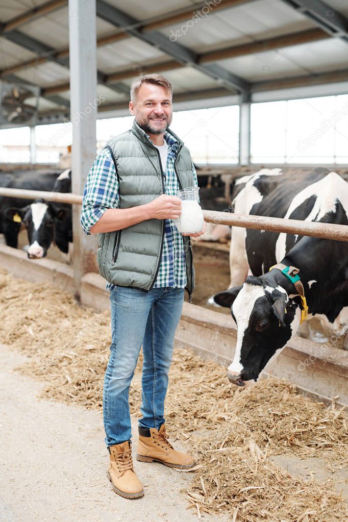 Healthy young farmer with jug of milk looking at you while standing inside dairy farm by cowshed
